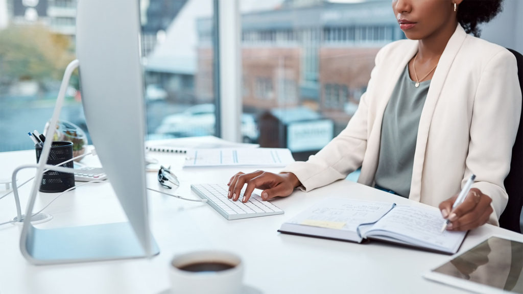 uma mulher negra com cabelos presos usando um computador e escrevendo no seu caderno de anotações.