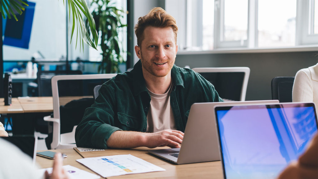 Homem de cabelos ruivos sorrindo em frente ao notebook.