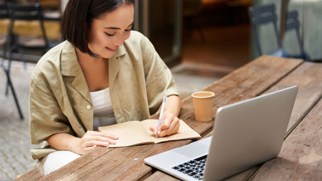 Uma mulher oriental de cabelos curtos escrevendo em um caderno em frente ao notebook. Vamos apresentar a importância das atualizações de softwares e algumas dicas para você iniciá-las agora mesmo.