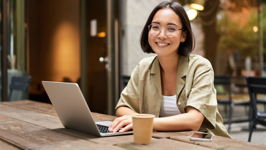 Mulher oriental de óculos e cabelo curto sorrindo em frente so notebook. Saiba por onde começar as atualizações de softwares