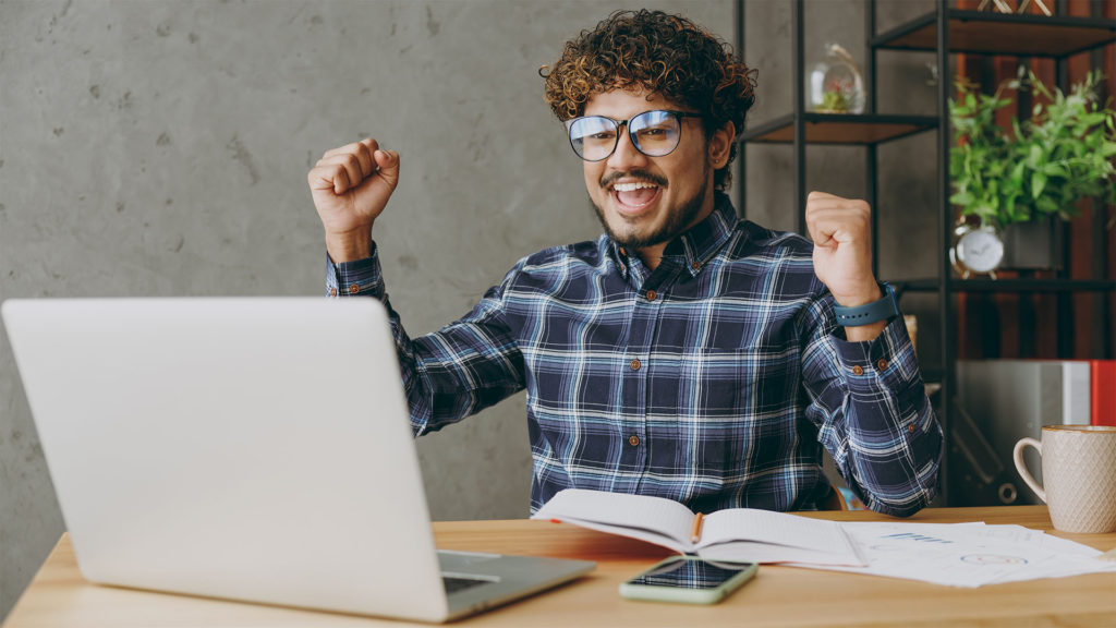 Homem em frente ao notebook sorrindo com os braços levantados, num gesto de comemoração