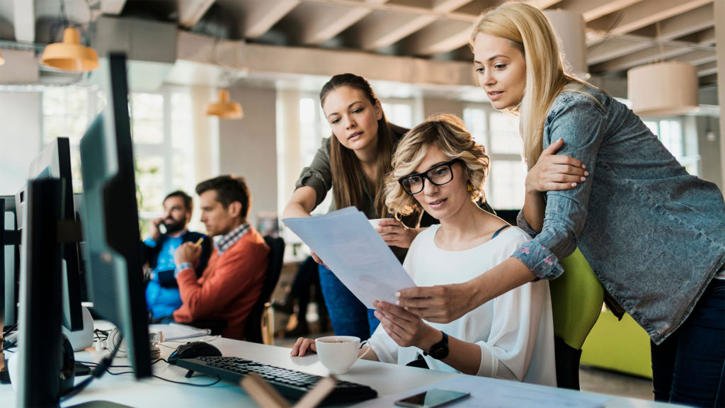 Três mulheres em frente ao computador olhando para uma folha de papel com expressões apreensivas. Por que será que os silos de dados são problemáticos?