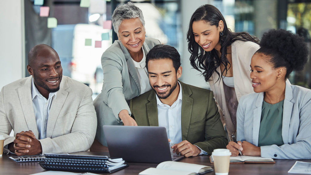 Equipe sorridente de trabalho reunida em frente ao notebook enquanto uma mulher aponta para a tela do computador portátil. Você conhece os benefícios da IA para empresas?