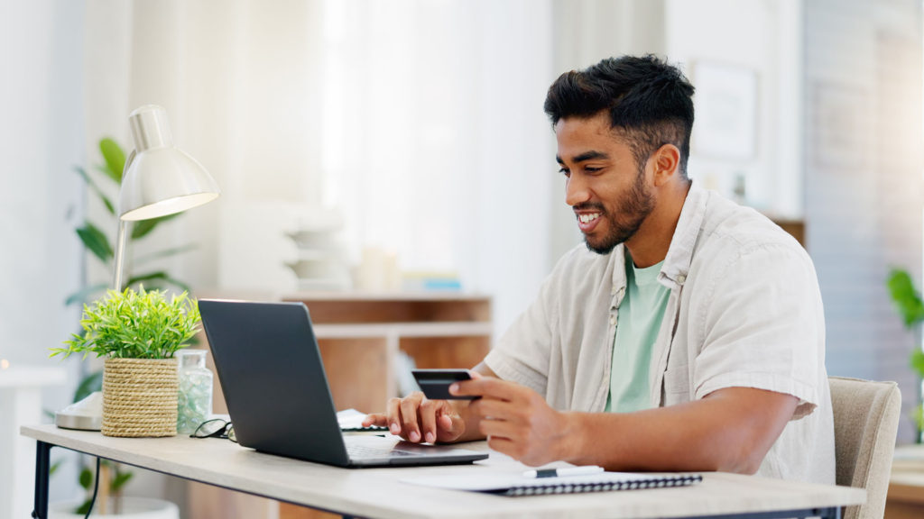 Homem sorrindo em frente ao notebook com cartão bancário nas mãos.