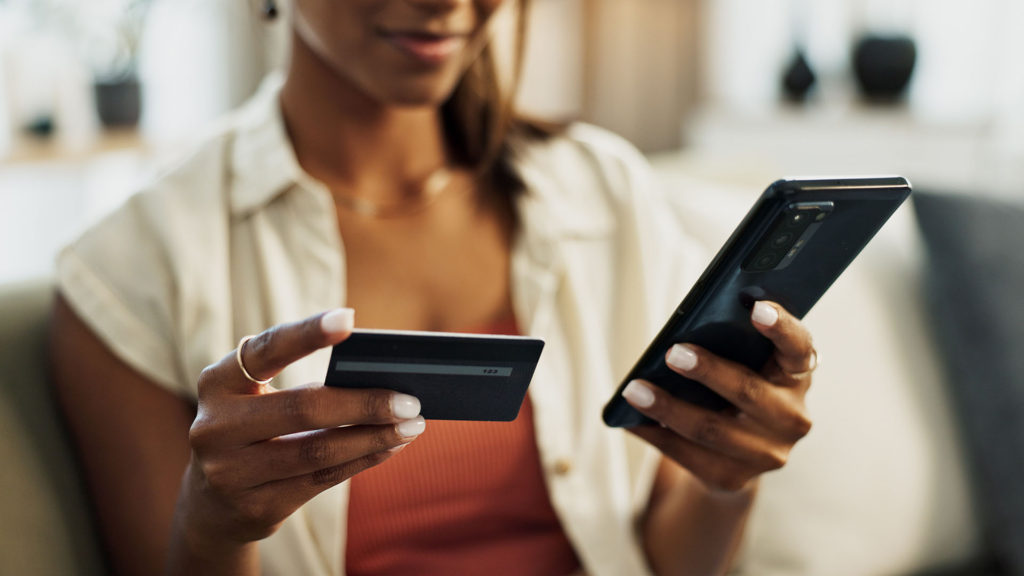 Mulher segurando o cartão de credito em uma das mãos enquanto realiza compra pelo celular