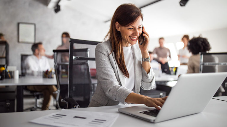 Mulher atendendo ao celugar enquanto usa o notebook. Investir em tecnologia é um passo essencial para atender as necessidades e expectativas do seu público, mas há alguns desafios em implantá-las.