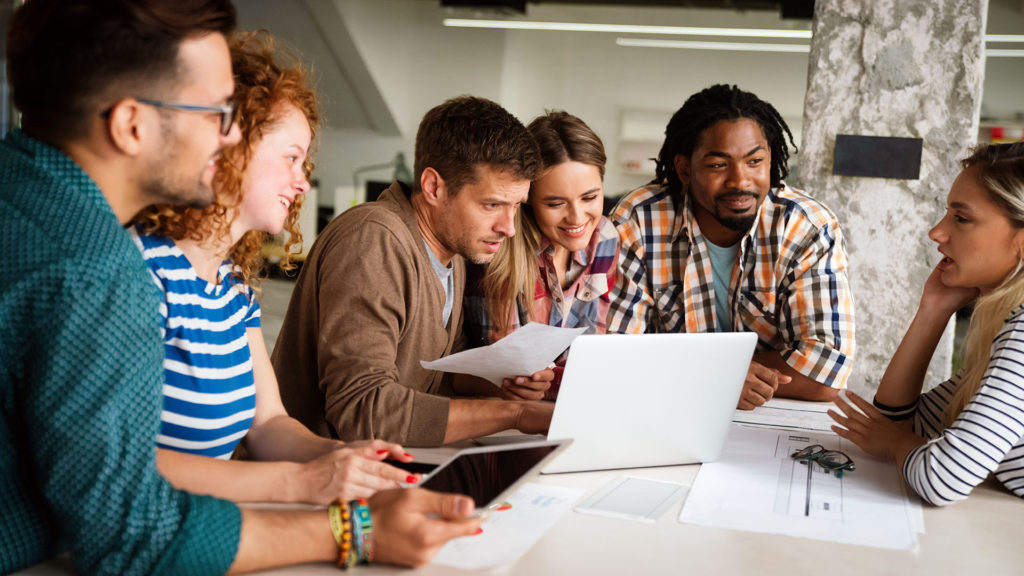 Equipe de trabalho de três homens e três mulheres usando um notebook sorrindo numa mesa de reunião. Você conhece os impactos da eficiência digital?