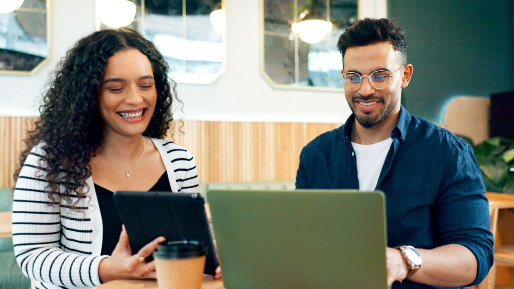 Dois colegas de trabalho, uma mulher e um home sorridentes, em frente ao notebook. Os benefícios da gestão de TI traz benefícios para segurança, produtividade, inovação e competitividade.