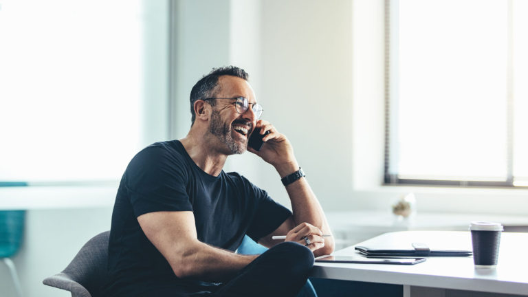 Homem sorrindo usando um celular em frente ao notebook. Saiba como a transformação digital nas empresas pode trazer muitos benefícios.